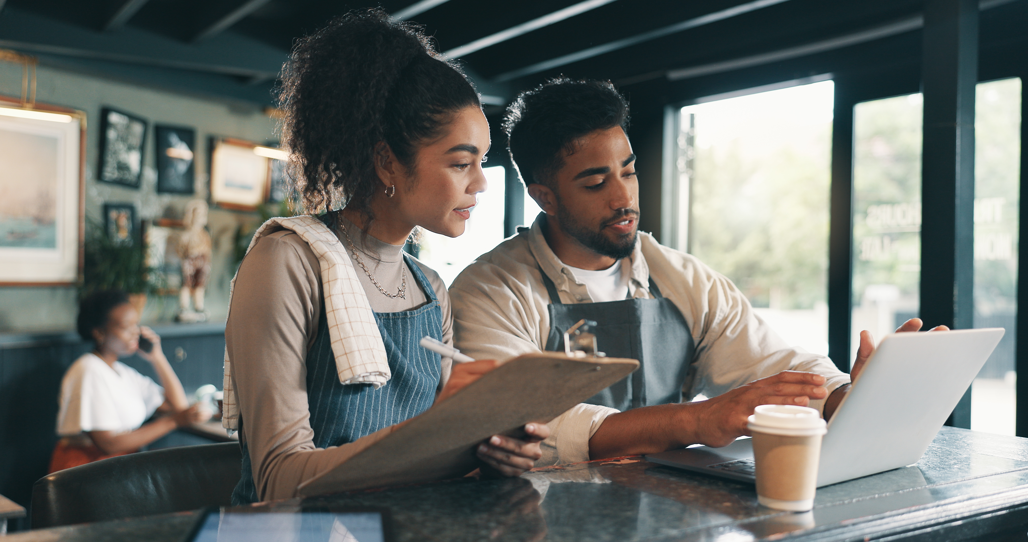 A small business owner and their first employee collaborating over a laptop together inside a cafe.